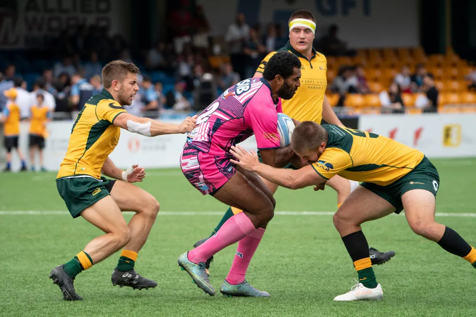 OSOB player in pink is tackled around the waist by two Classic Wallabies in green and gold beside a banner
