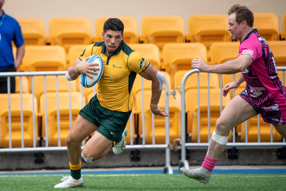 Classic Wallabies winger in green and gold runs the ball along a railing of empty yellow seats, an OSOB chaser behind