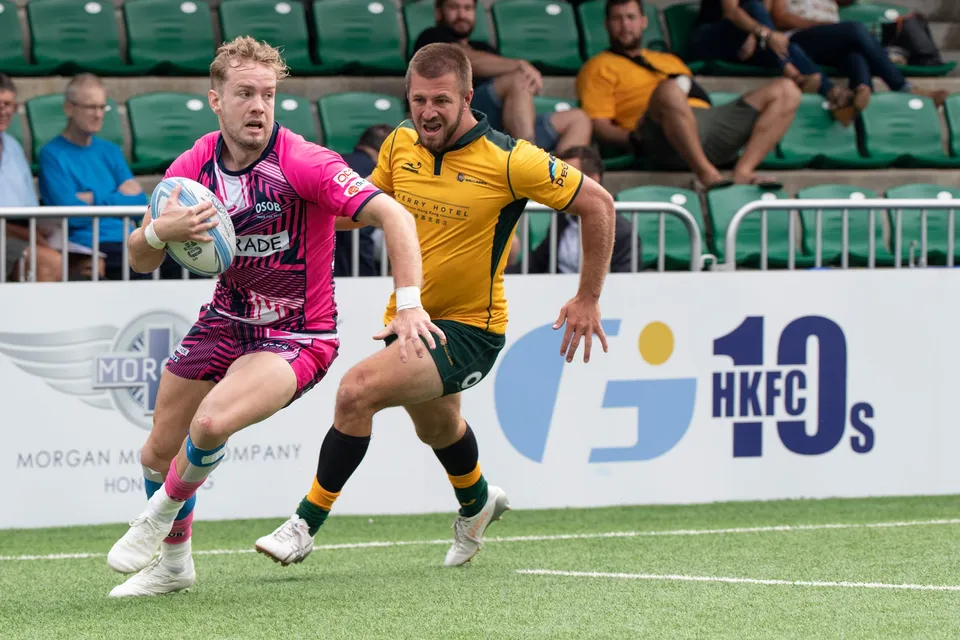 Blond OSOB ball carrier sprints before the HKFC 10s banner with a Classic Wallabies defender alongside