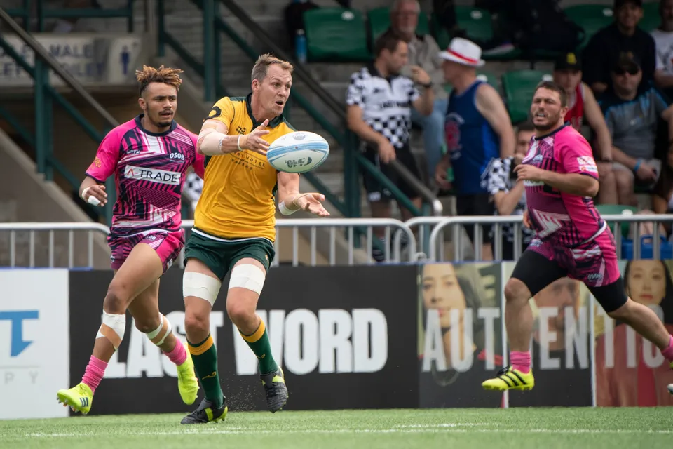 Classic Wallabies player in gold throws a pass as OSOB players in pink run in from both sides, fans in the stand