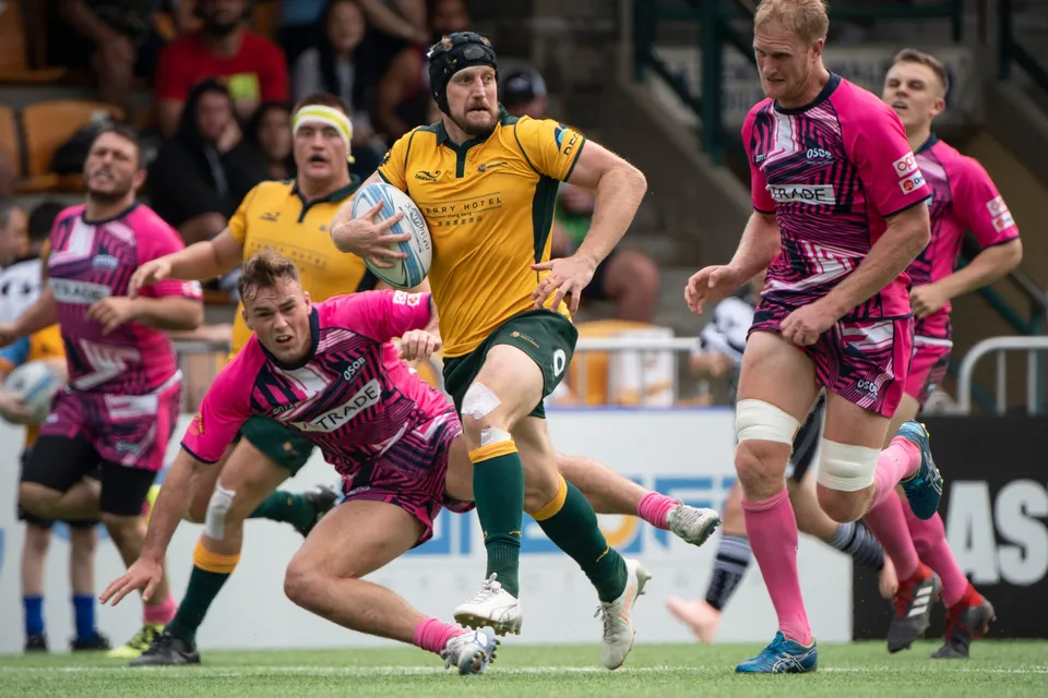 Wallabies ball carrier in a scrum cap breaks away as an OSOB player in pink dives at his feet