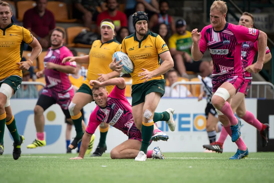 Wallabies runner in a scrum cap carries the ball forward as an OSOB tackler slides along the grass below him