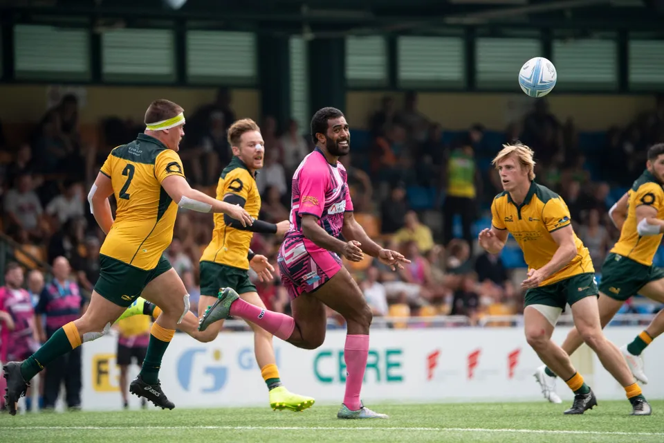 OSOB player in pink runs with arms out as the ball sails overhead, Classic Wallabies players looking up behind
