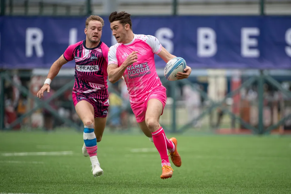 Waterboys player in plain pink runs with the ball past an OSOB team-mate in a patterned shirt, banner behind