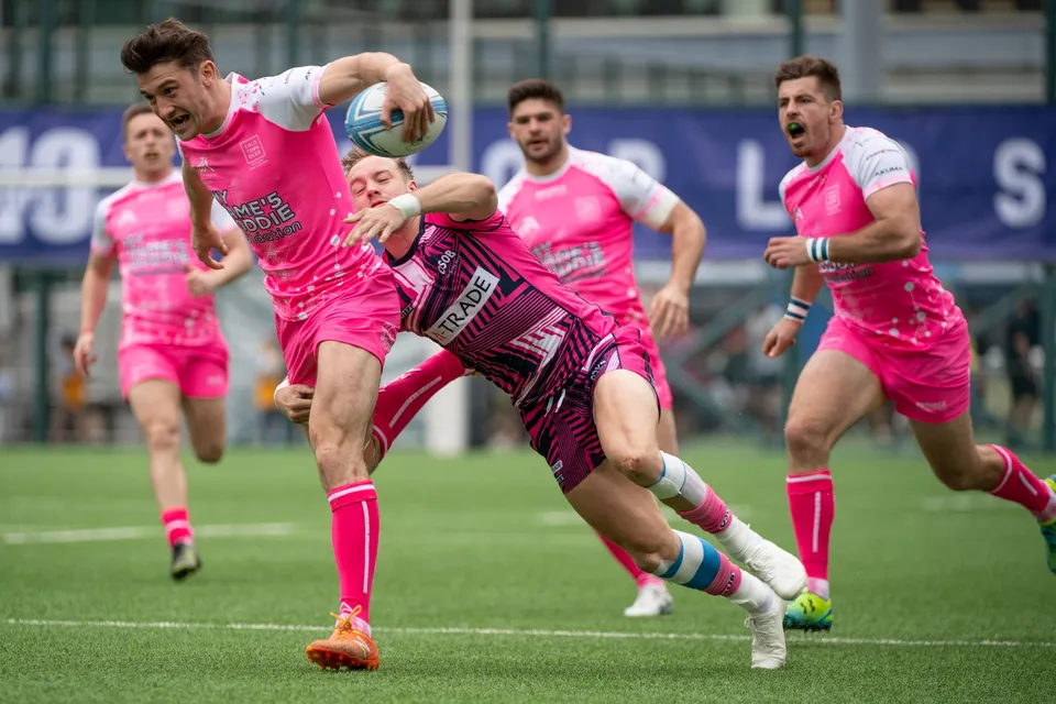 Projecx Waterboys ball carrier in plain pink is tackled around the waist by an OSOB player in a patterned shirt