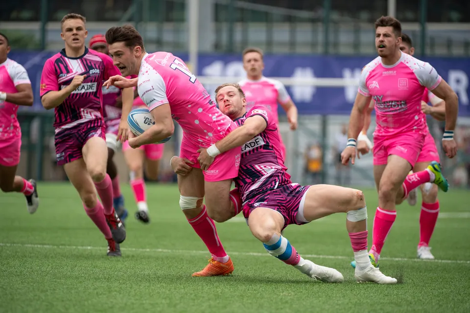 OSOB defender in a patterned shirt grabs a Waterboys ball carrier in plain pink as pink-shirted team-mates follow up