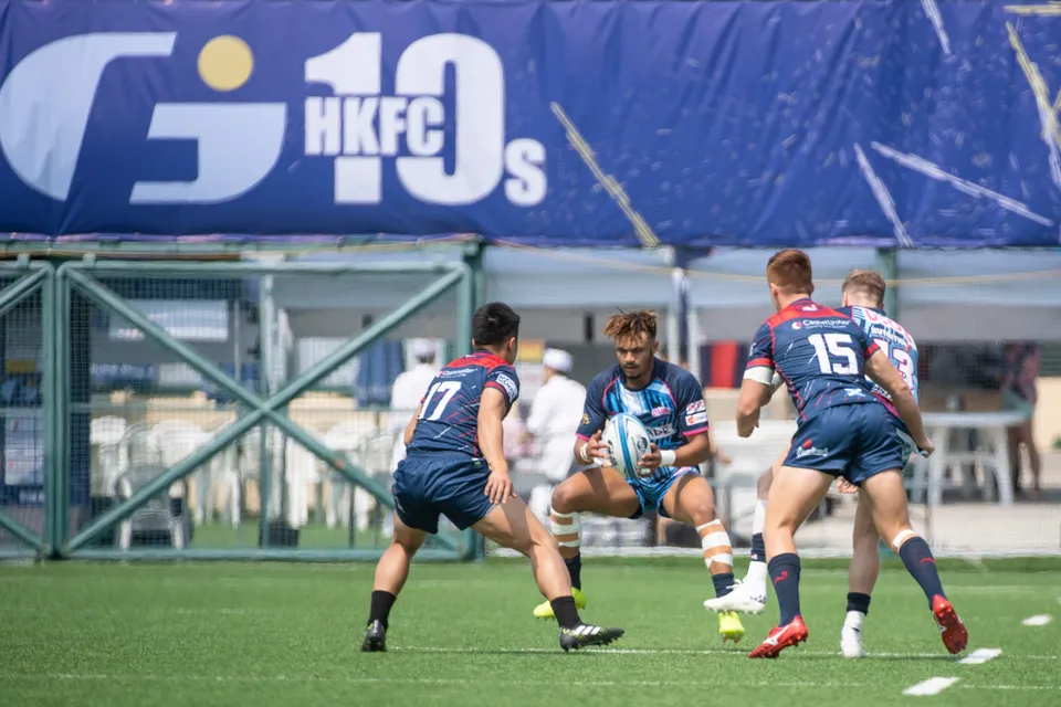 OSOB player in light blue holds the ball facing two navy-kit Scottish Exiles defenders below an HKFC 10s banner