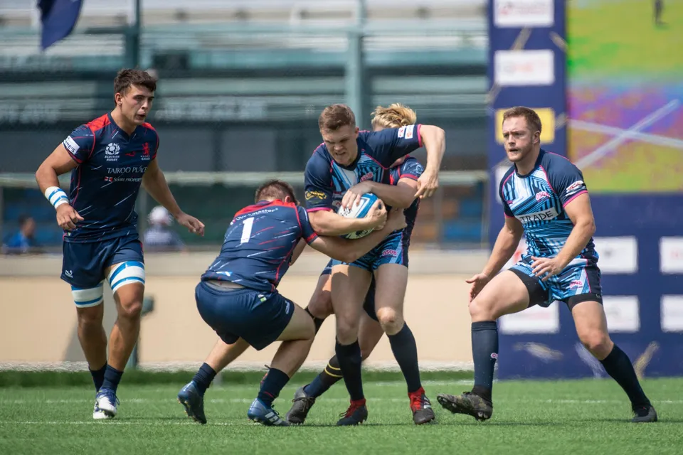Ball carrier is gripped by a low tackler against Taikoo Place Scottish Exiles while players wait either side