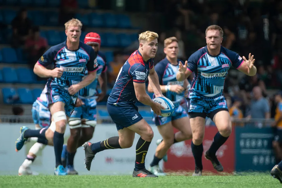 Blond Scottish Exiles player in navy carries the ball as a light blue OSOB defender raises his hands beside him