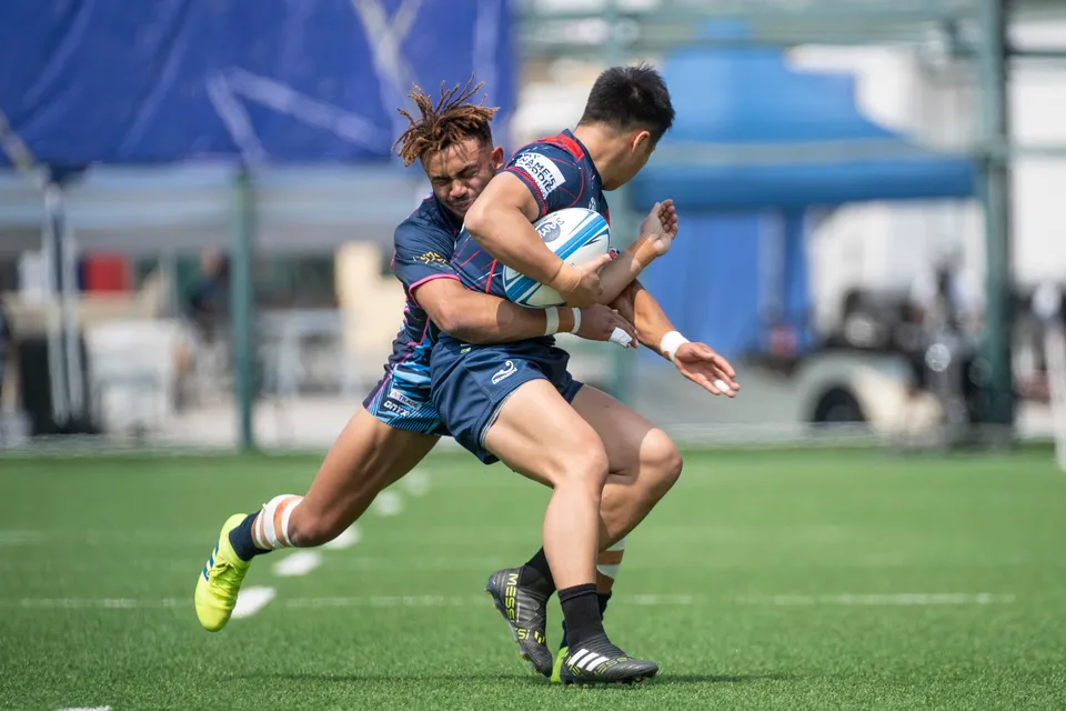 Navy-kit ball carrier is grabbed round the body by a light blue OSOB tackler on artificial turf