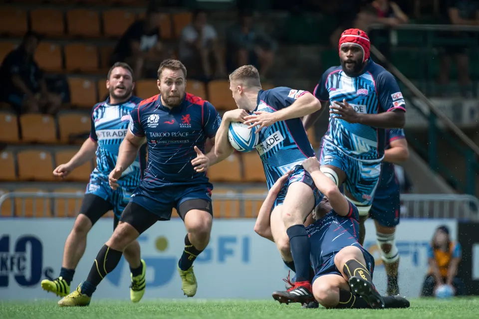 Scottish Exiles ball carrier in navy is tackled low by an OSOB player as a headgear-wearing team-mate arrives behind