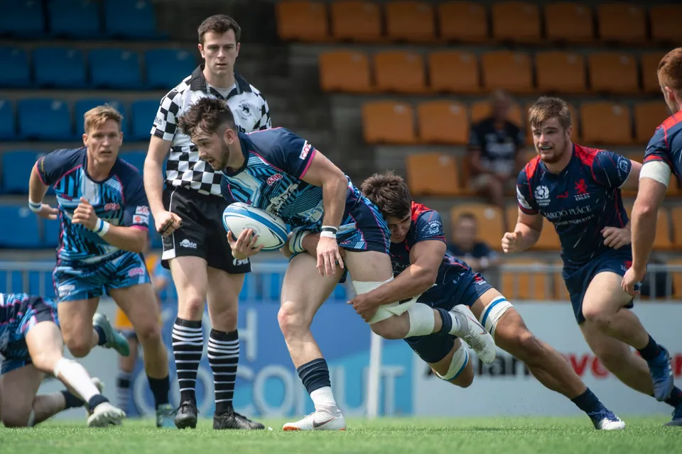 Ball carrier in light blue stripes is tackled low by a navy defender while the referee and teammates look on in 2019