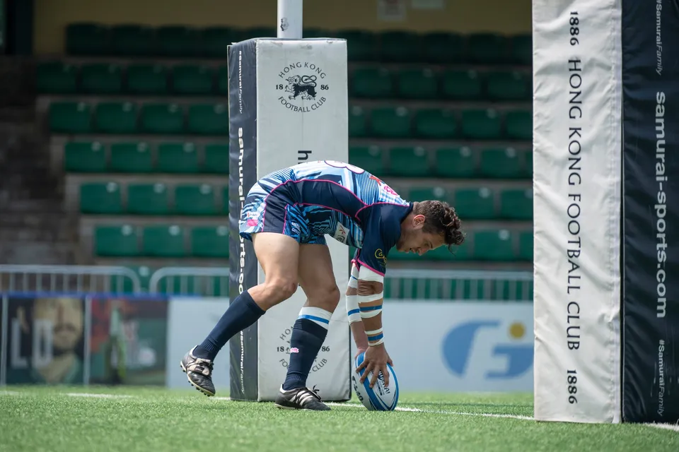 Player in blue touches the ball down beside a padded Hong Kong Football Club post in front of green stands, 2019