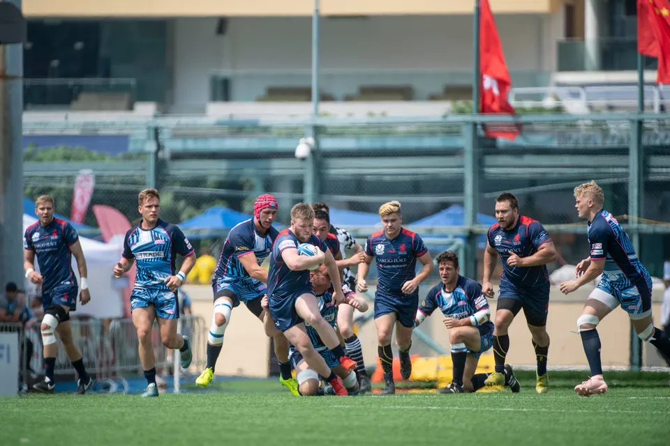 Navy-shirted runner drives forward with the ball as teammates in navy and light blue back him up in 2019 Exiles game