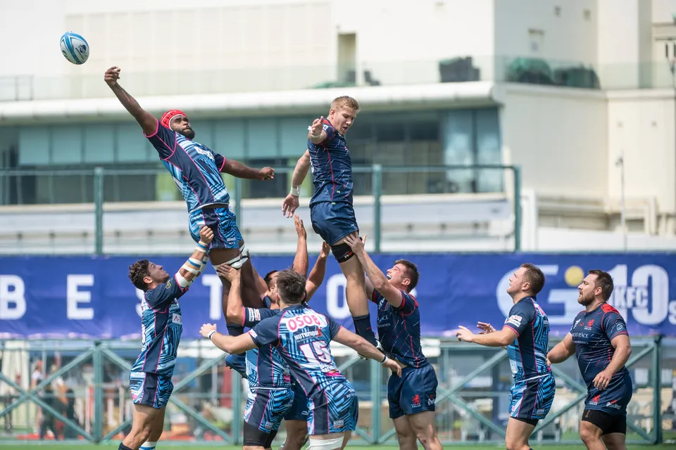 Line-out jumper in blue is lifted high with arm stretched for the ball beneath a blue HKFC 10s banner, 2019
