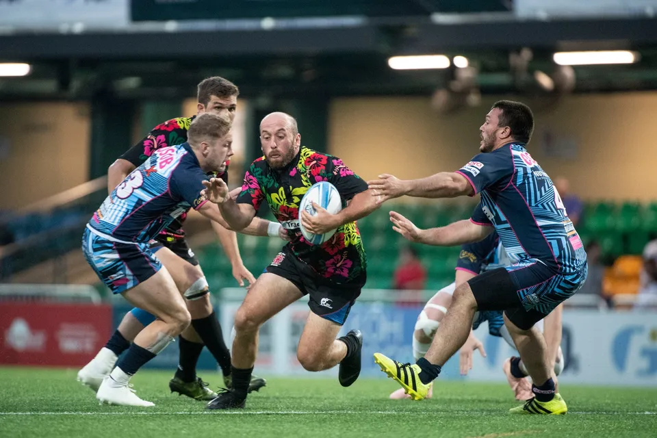 Ball carrier in a black floral shirt drives between two blue-patterned defenders in the 2019 YCAC match
