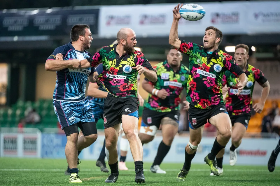 Player in a black floral shirt passes the ball off one hand while a blue-striped opponent closes in, 2019 YCAC