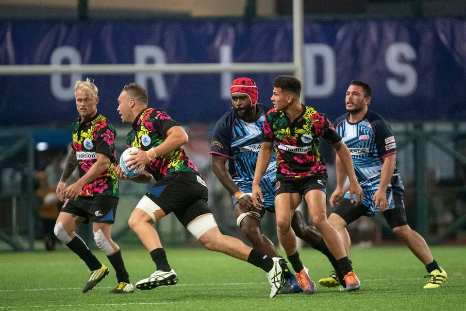 Floral-shirted ball carrier runs in open play with four players around him in front of a blue banner, 2019 YCAC