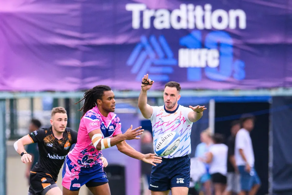 OSOB player in pink and blue flicks the ball away beside a referee raising an arm, Ashbury Tropics defender close behind