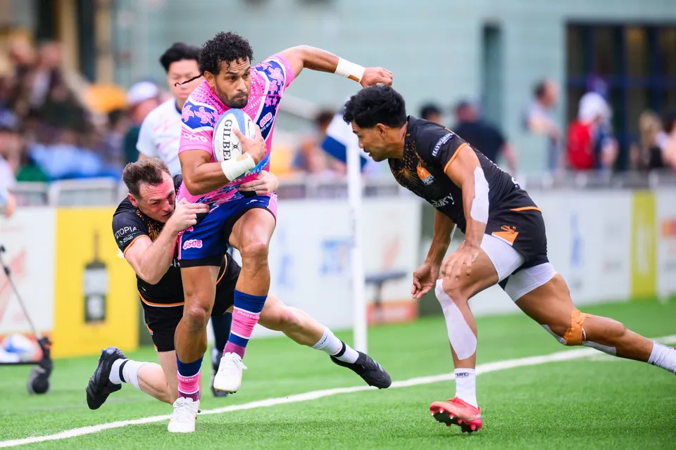 OSOB ball carrier is wrapped by two Ashbury Tropics defenders in black as a third closes in from the right