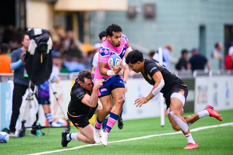 OSOB ball carrier twists in a tackle as an Ashbury Tropics defender slides along the grass and a camera operator watches