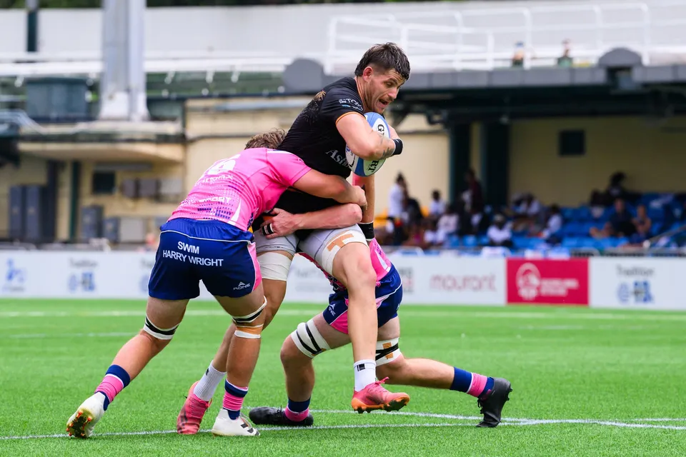 Ashbury Tropics ball carrier is held up by two OSOB defenders in a tackle in front of the advertising boards