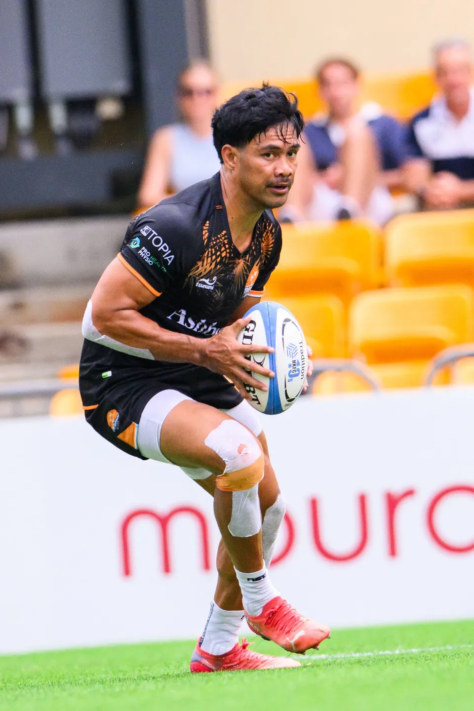 Ashbury Tropics player in black with taped knee and orange boots holds the ball in front of a yellow stand