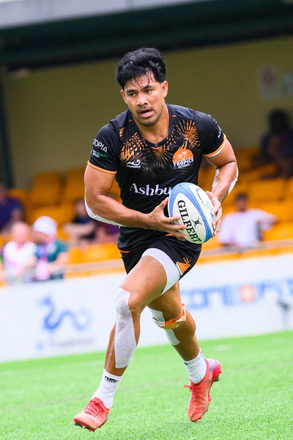 Ashbury Tropics player in black with orange boots runs with the ball in two hands in front of yellow stands