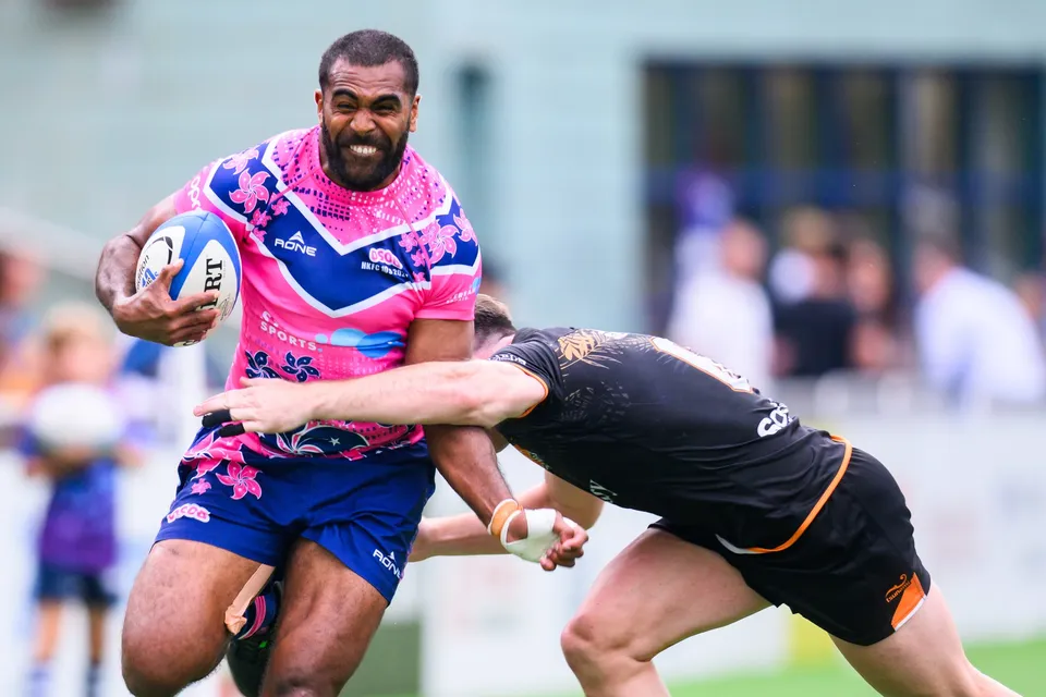 Smiling OSOB ball carrier is clutched at the waist by an Ashbury Tropics defender in black while running with the ball