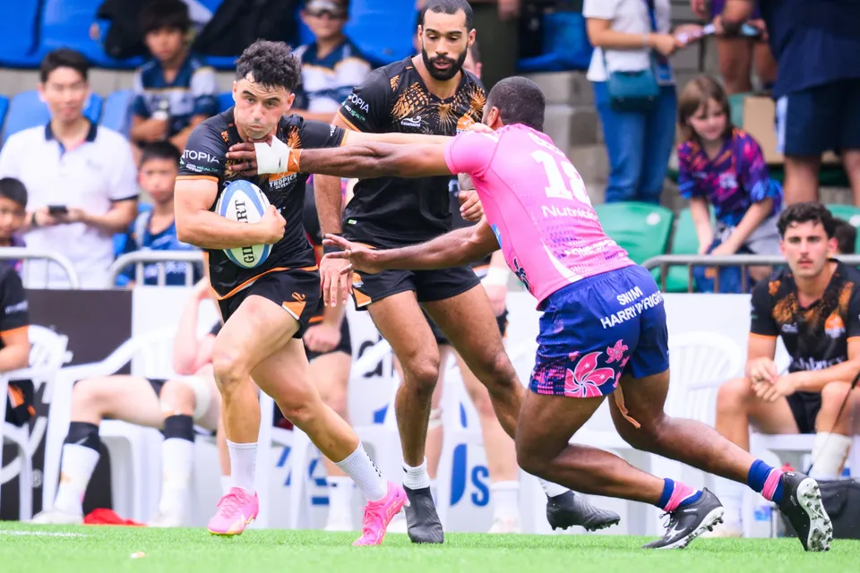 Ashbury Tropics ball carrier runs past an outstretched OSOB defender in front of a stand of spectators
