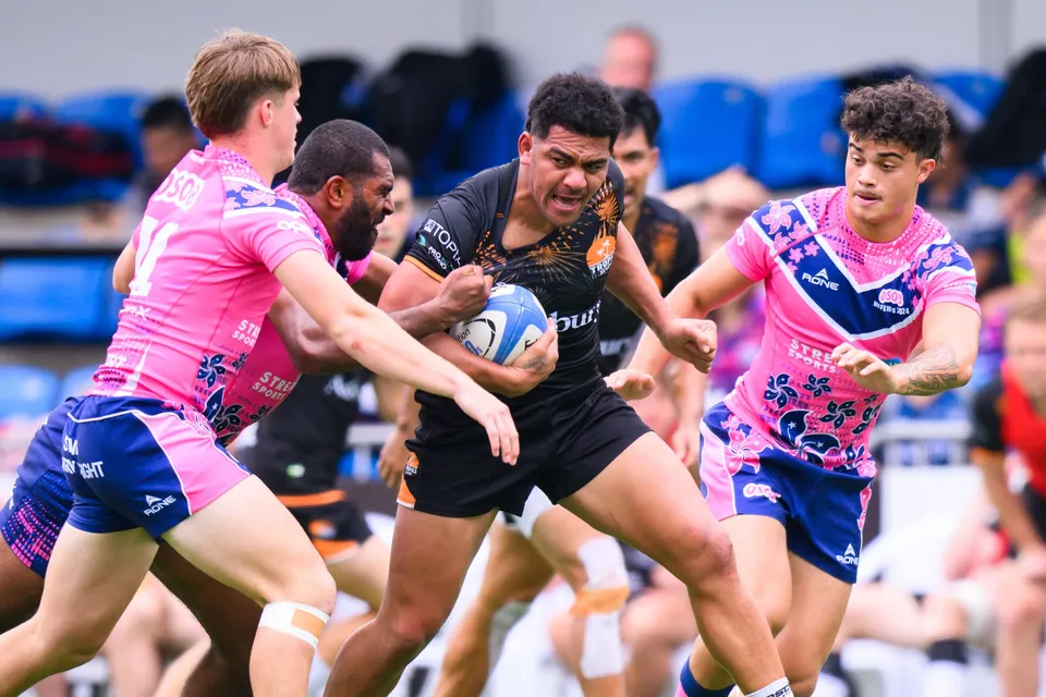Ashbury Tropics runner clutching the ball is hit by an OSOB tackler from behind, blue seats behind