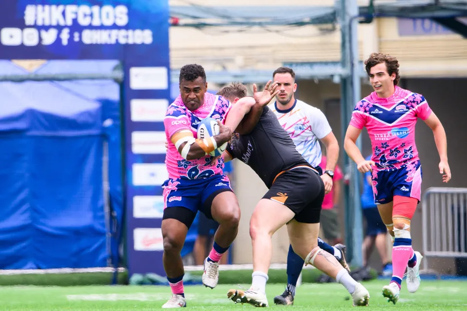 OSOB ball carrier shields the ball as an Ashbury Tropics defender wraps him, in front of a HKFC 10s banner