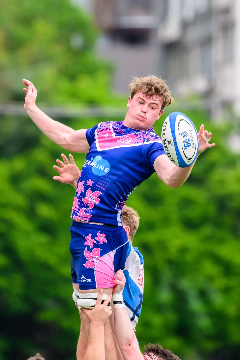 OSOB line-out jumper, lifted by teammates, stretches both arms to catch the ball against green trees