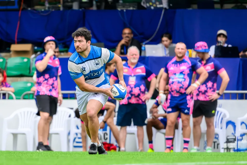 HKFC Natixis player in light blue hoops looks to pass while OSOB substitutes in pink watch from the sideline