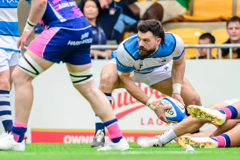 HKFC Natixis player with a beard crouches low, ball near the grass, while OSOB legs fill the foreground
