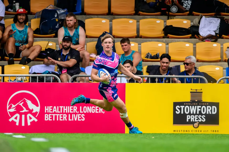 OSOB runner carries the ball near the touchline under AIA and Stowford Press boards as spectators look on