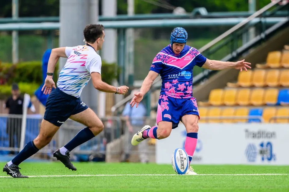 OSOB kicker in a blue scrum cap swings his boot at the ball as an opponent in a white shirt charges