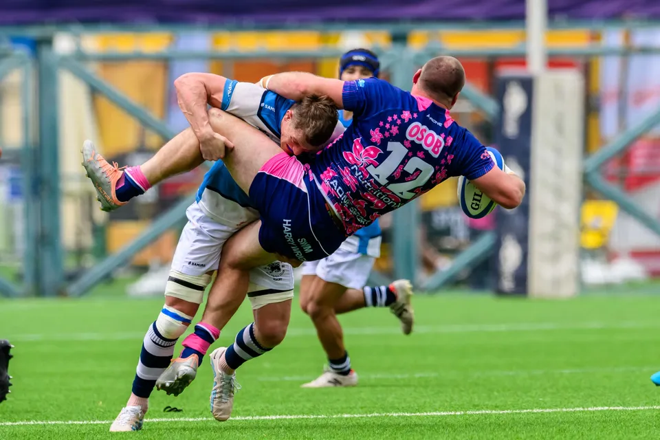 OSOB number 12 is upended horizontally in a tackle by an HKFC Natixis player, the ball at his right hand