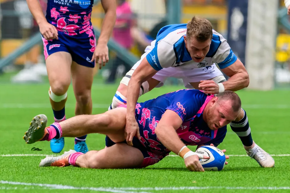 OSOB player stretches along the grass, ball tucked under his arm, with an HKFC Natixis defender leaning over him