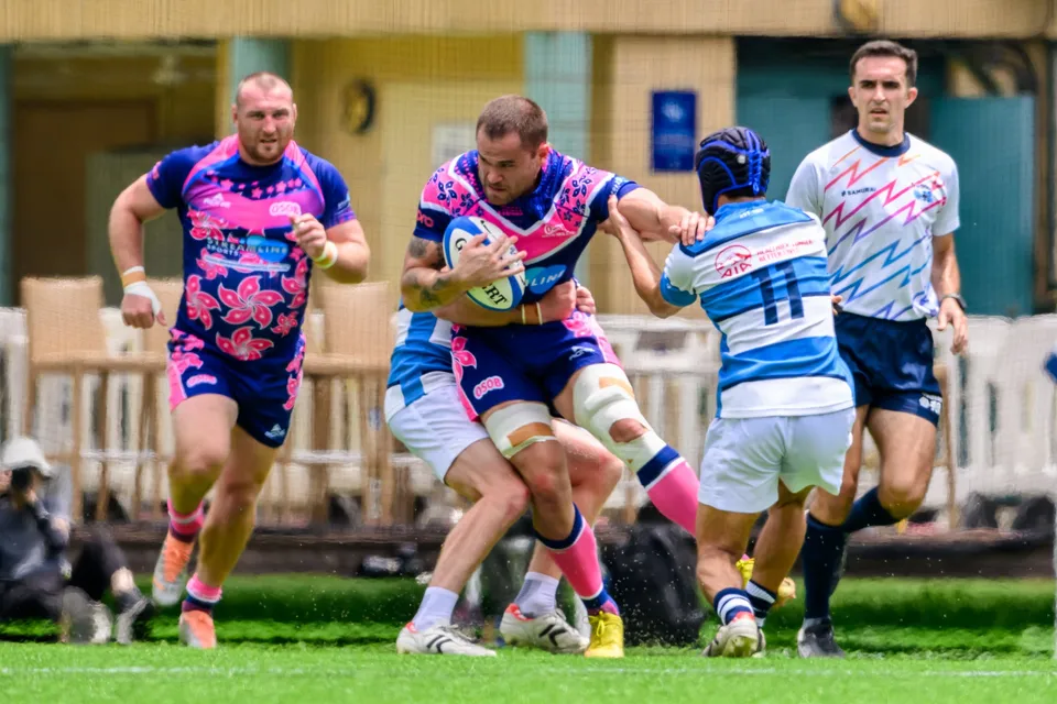 OSOB ball carrier is tackled by an HKFC Natixis player in a scrum cap as an OSOB teammate runs up in support