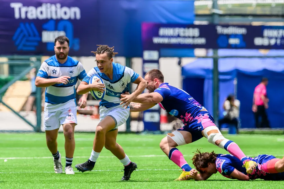 HKFC Natixis runner holds the ball in both hands and steps past a diving OSOB tackler under a Tradition banner