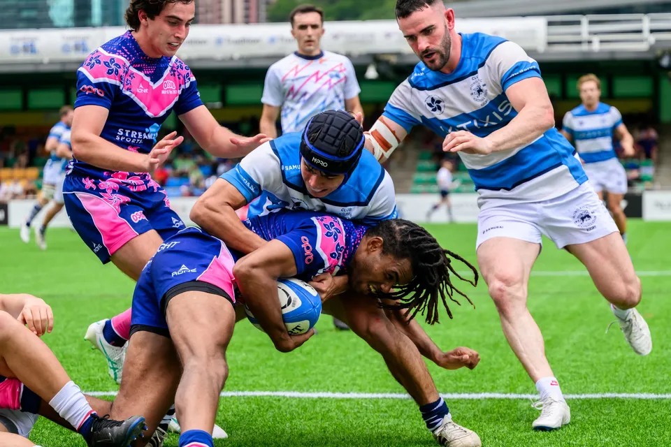 OSOB ball carrier drives low through a tackle by an HKFC Natixis player in a scrum cap as teammates close in