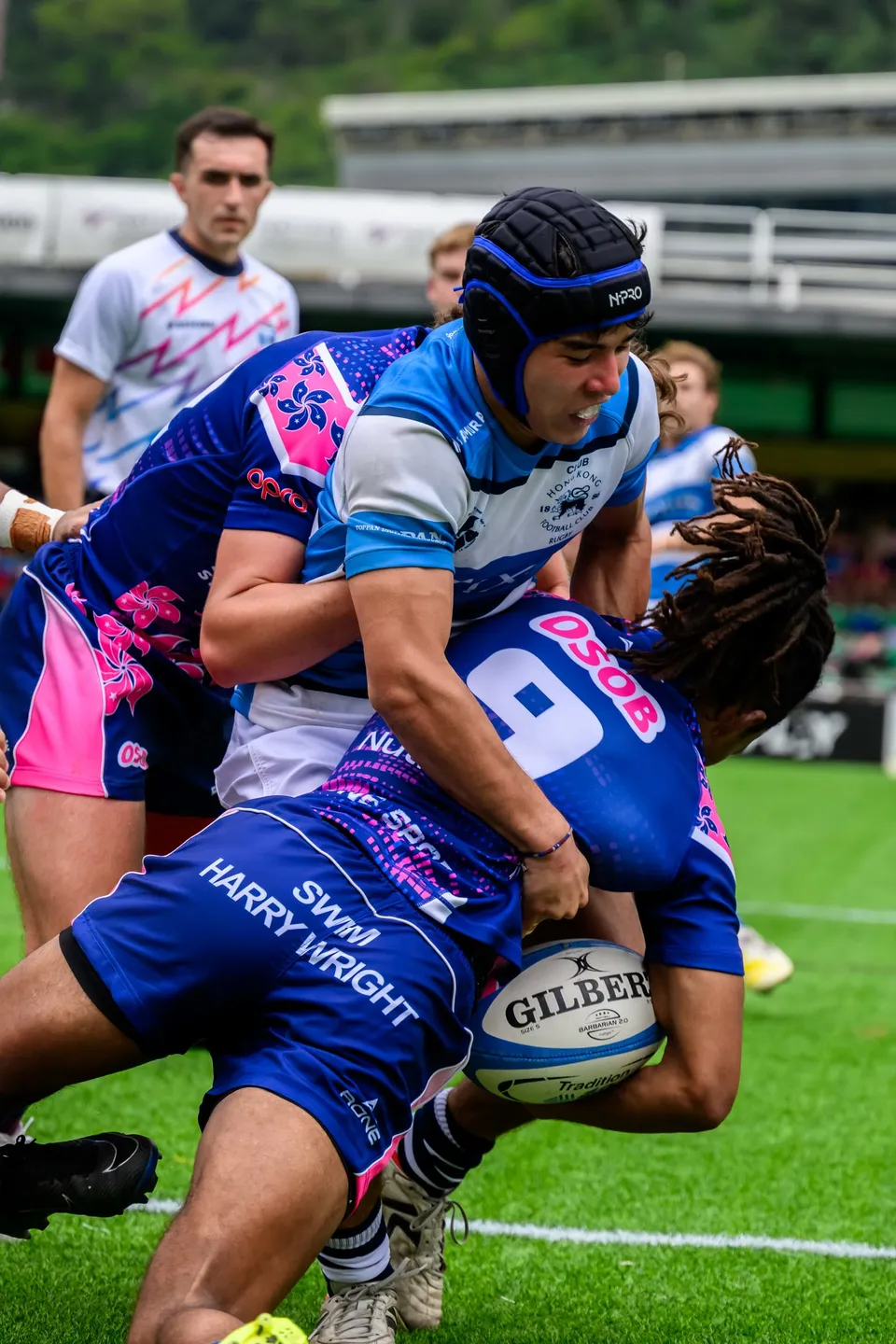 OSOB number 9 holds the ball in a tackle by an HKFC Natixis player in a scrum cap, referee behind