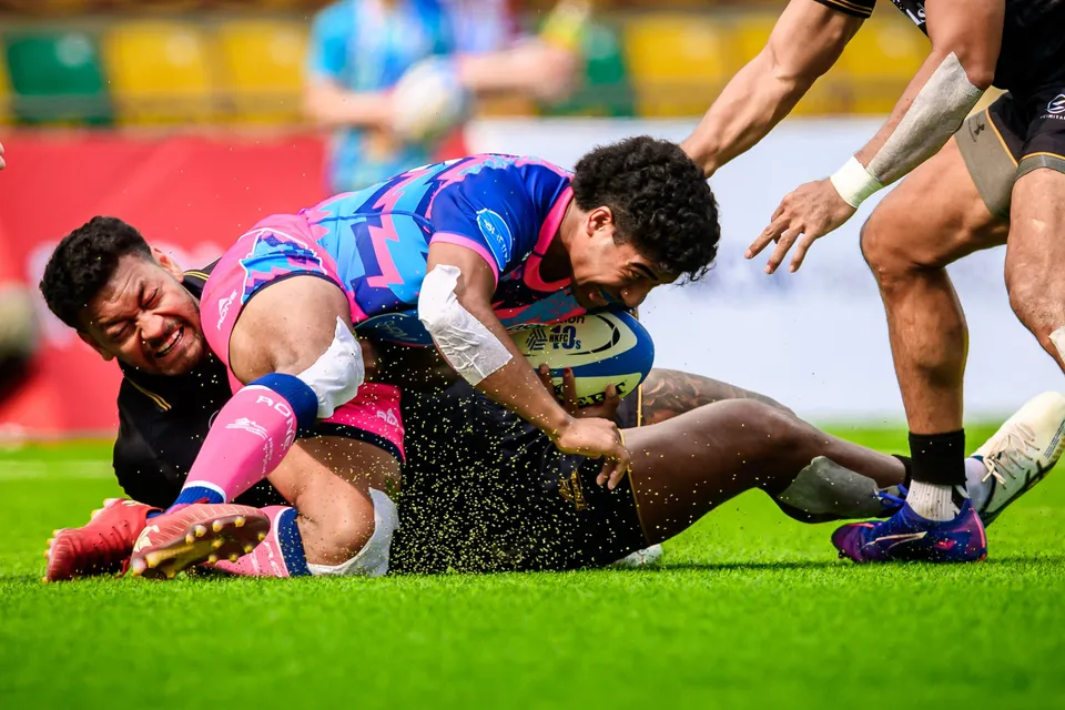 OSOB player in pink falls forward on the grass with the ball over an Ashbury Tropics player lying beneath him