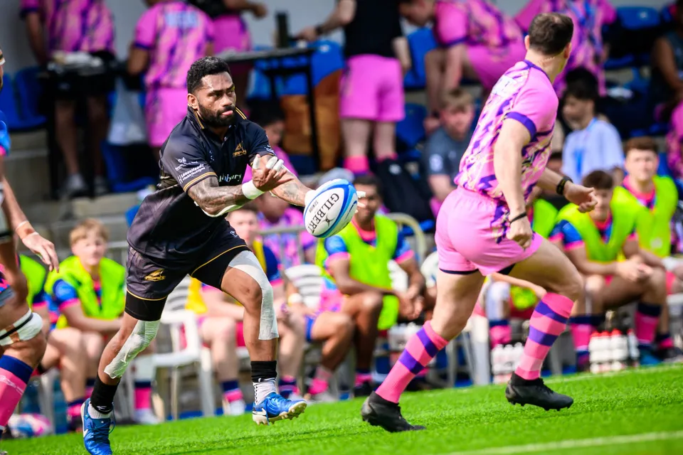 Ashbury Tropics player in black passes the ball on the run while an OSOB player in pink chases, with spectators behind