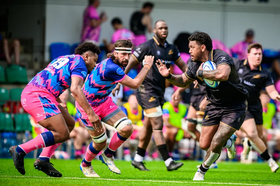 Ashbury Tropics ball carrier in black runs at two OSOB defenders in pink and blue, spectators behind
