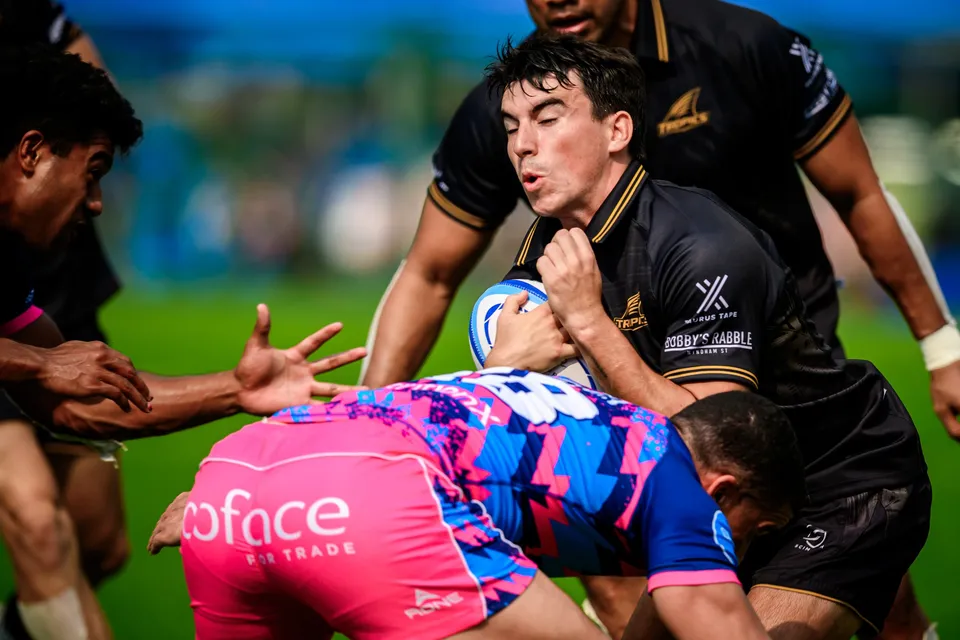 Close view of a Tropics player in black clutching the ball as an OSOB tackler in pink wraps him low