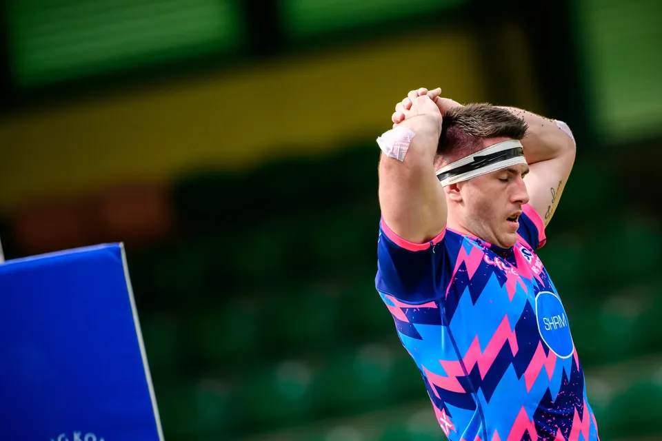 OSOB player in pink and blue shirt and headband stands with both hands on his head, blue board behind