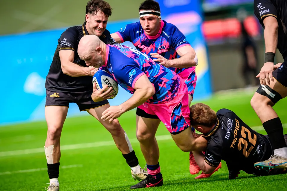 Bald OSOB ball carrier is wrapped up by two Tropics defenders in black, another opponent on the ground