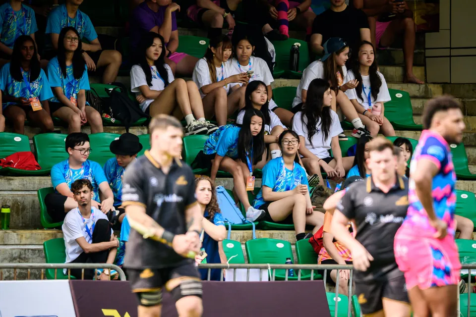 Young spectators in blue and white T-shirts watch from the stand as OSOB and Tropics players stand in front